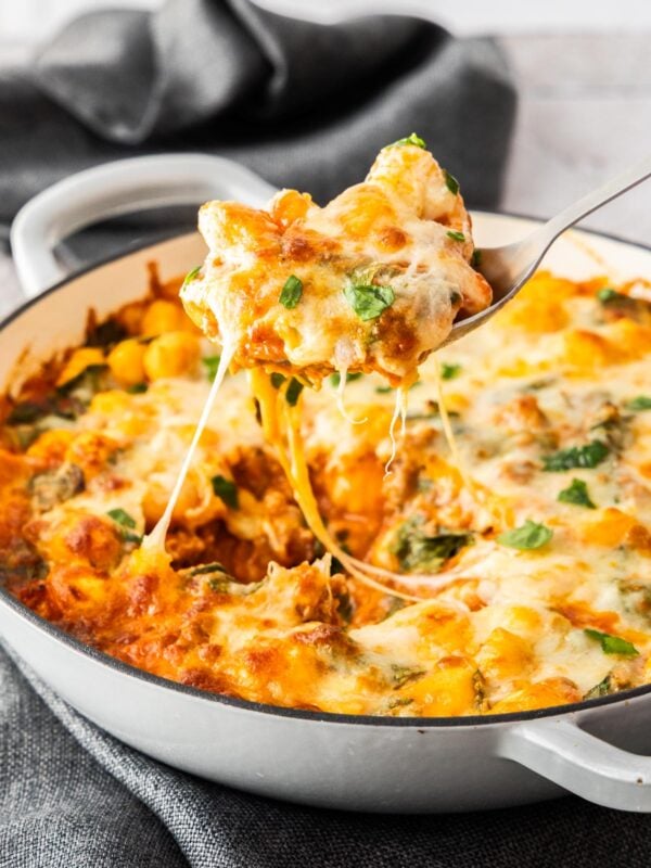 a spoon holding a scoop of gnocchi casserole over a baking dish, with cheese being pulled.