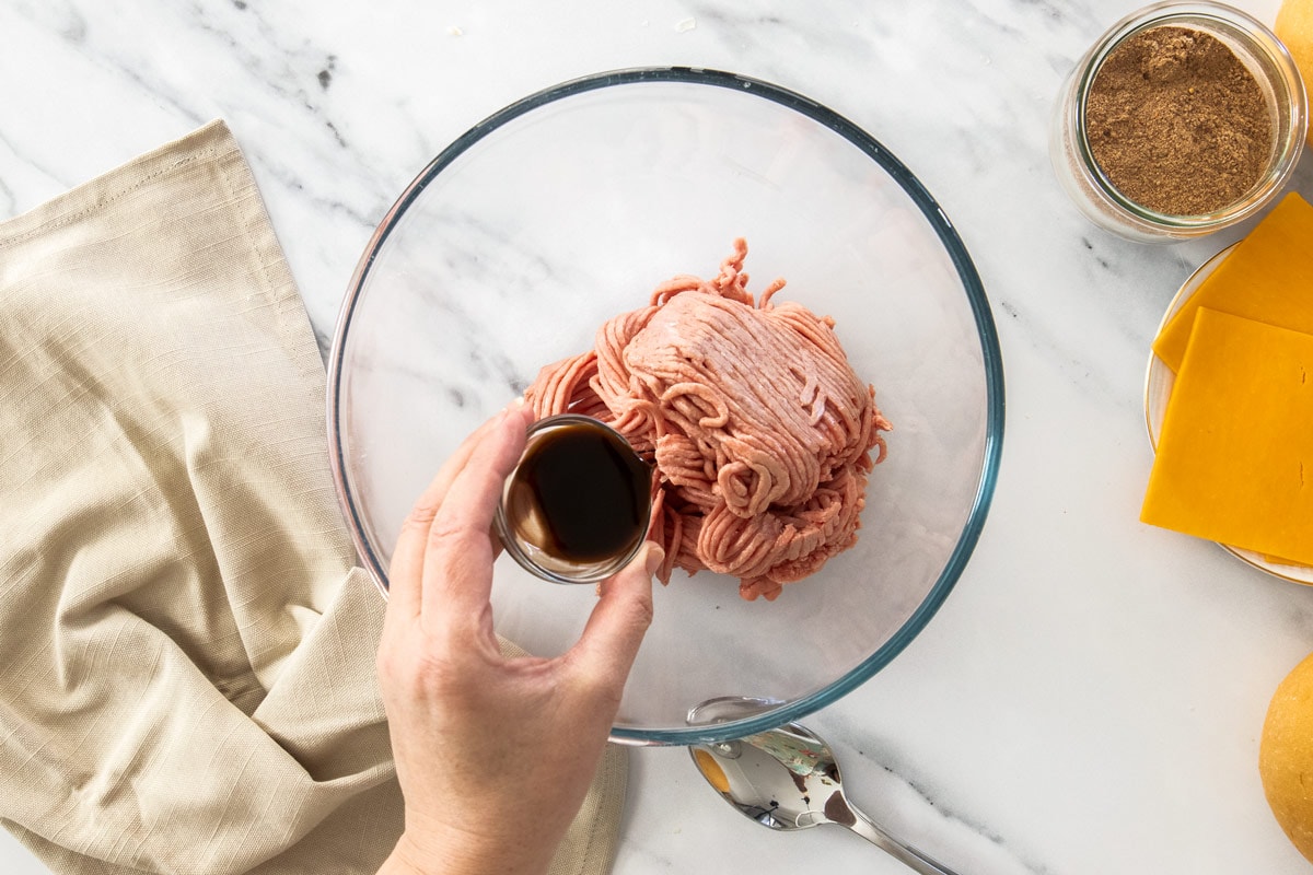 ground meat in a glass bowl with a jar of sauce being poured over it.