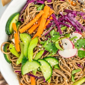 a white bowl filled with soba noodles, chopped colorful vegetables, and topped with black and white sesame seeds.