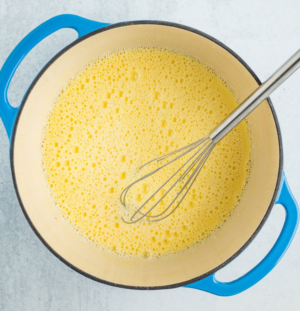 a blue dutch oven filled with dairy and a whisk.