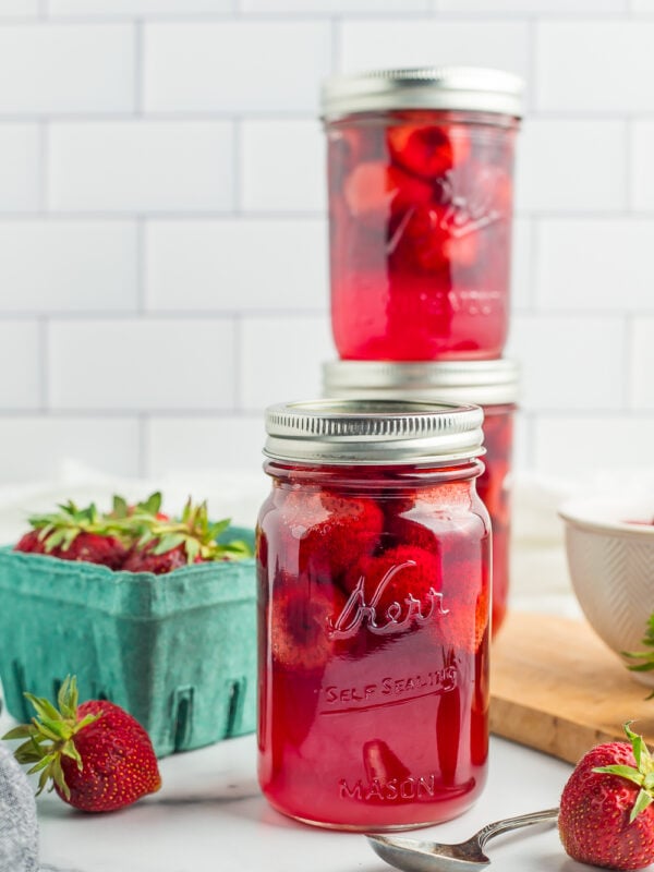 3 jars of strawberries on a marbled board with fresh strawberries in a small cardboard pint.