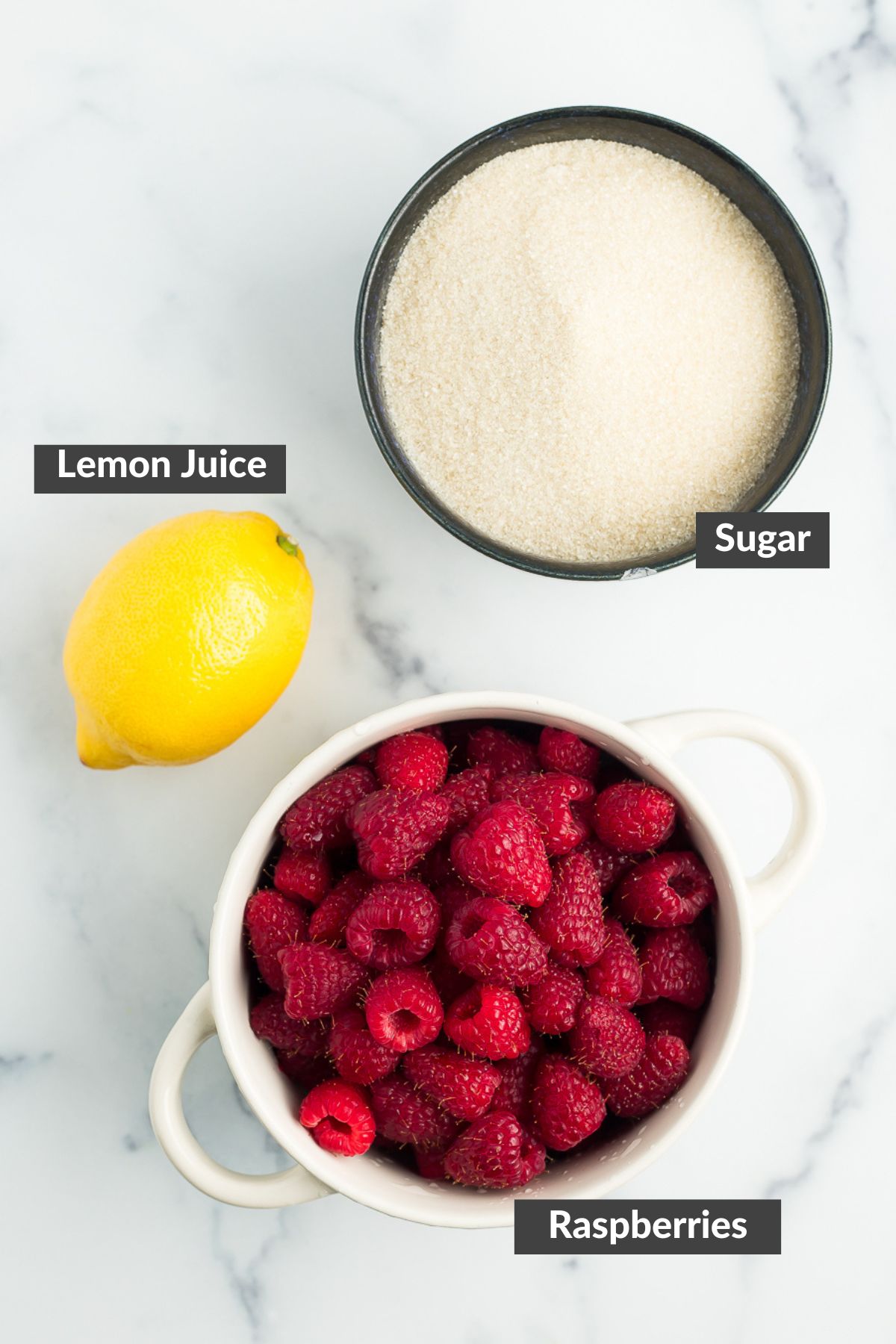 a lemon, a bowl of sugar, and a bowl of raspberries on a marbled board.