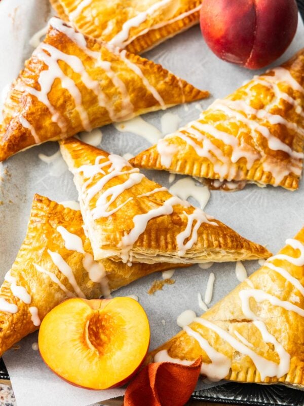 peach puff pastry turnovers on a baking sheet with sliced and whole peaches.