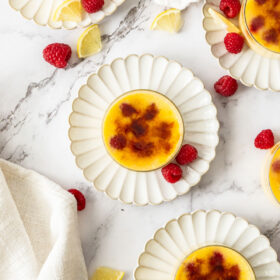 4 glass bowls of raspberry creme brulee on plates with fresh raspberries and lemon slices.