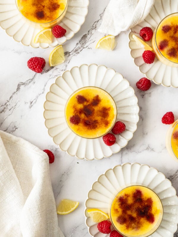 4 glass bowls of raspberry creme brulee on plates with fresh raspberries and lemon slices.