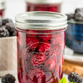 a jar of canned blackberries with a white and blue bowl of blackberries in the background.