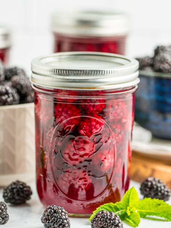 a jar of canned blackberries with a white and blue bowl of blackberries in the background.