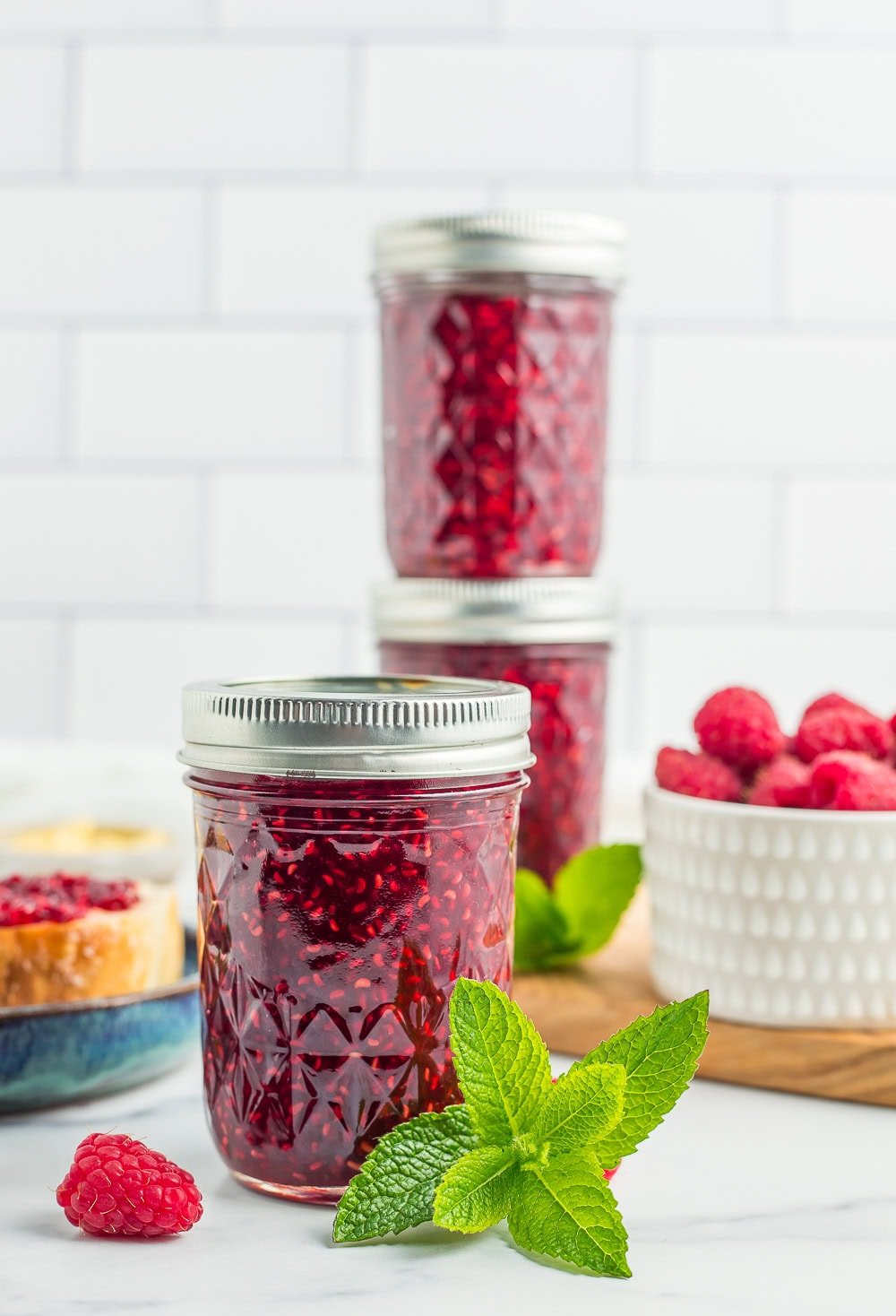 three jars of raspberry jam with fresh raspberries and a sprig of mint.