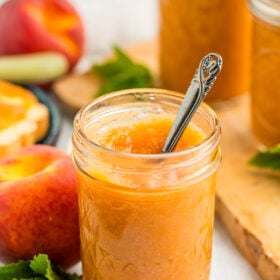 a jar of peach jam with a small spoon in it in front of other jars of jam, fresh peaches, and mint.