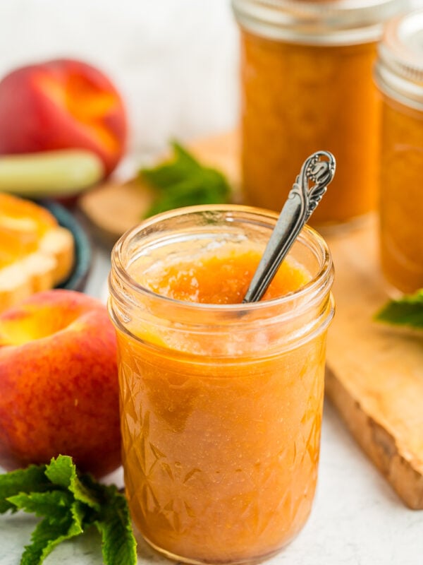 a jar of peach jam with a small spoon in it in front of other jars of jam, fresh peaches, and mint.