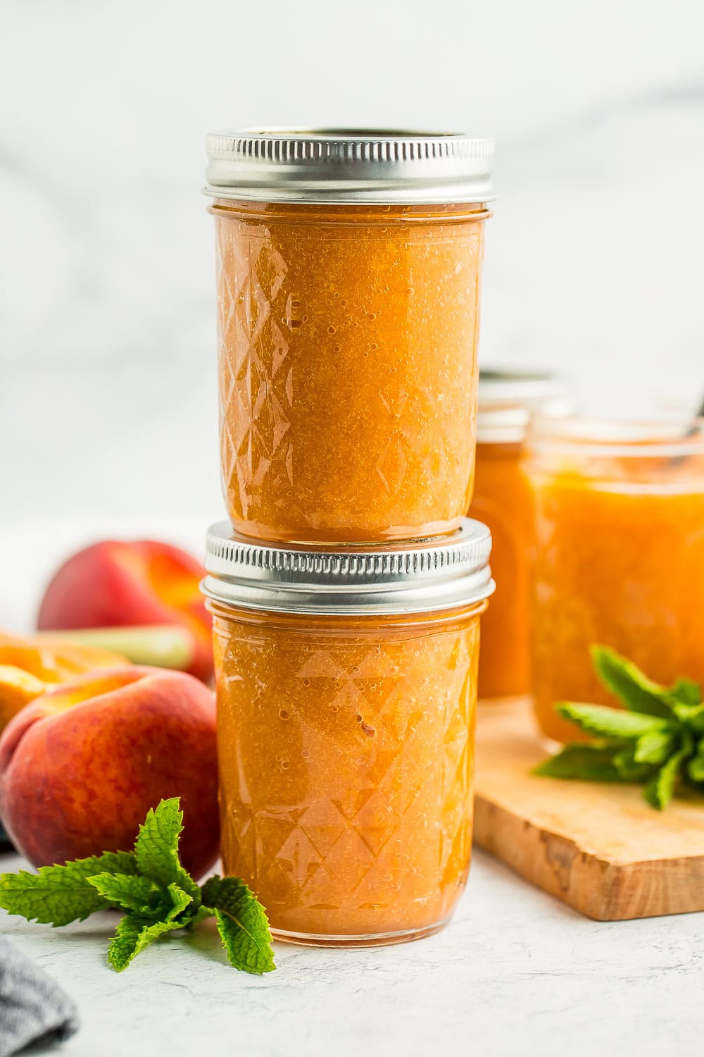 two jars of peach jam stacked on top of each other with fresh peaches and mint on a board.