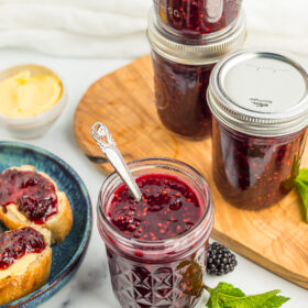 4 jars of blackberry jam with fresh blackberries and a sprig of mint.