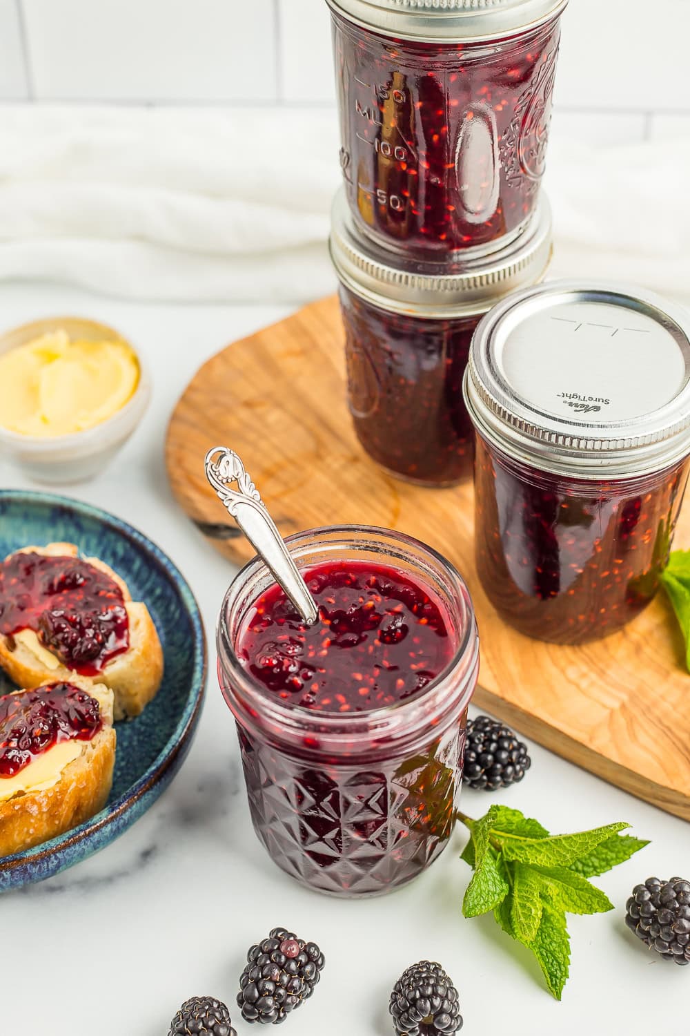 4 jars of blackberry jam with fresh blackberries and a sprig of mint.