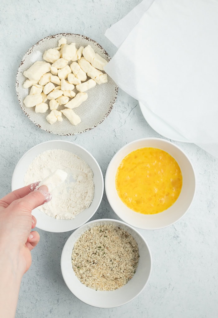 3 bowls for breading and a plate of cheese curds with a hand dipping one into flour.