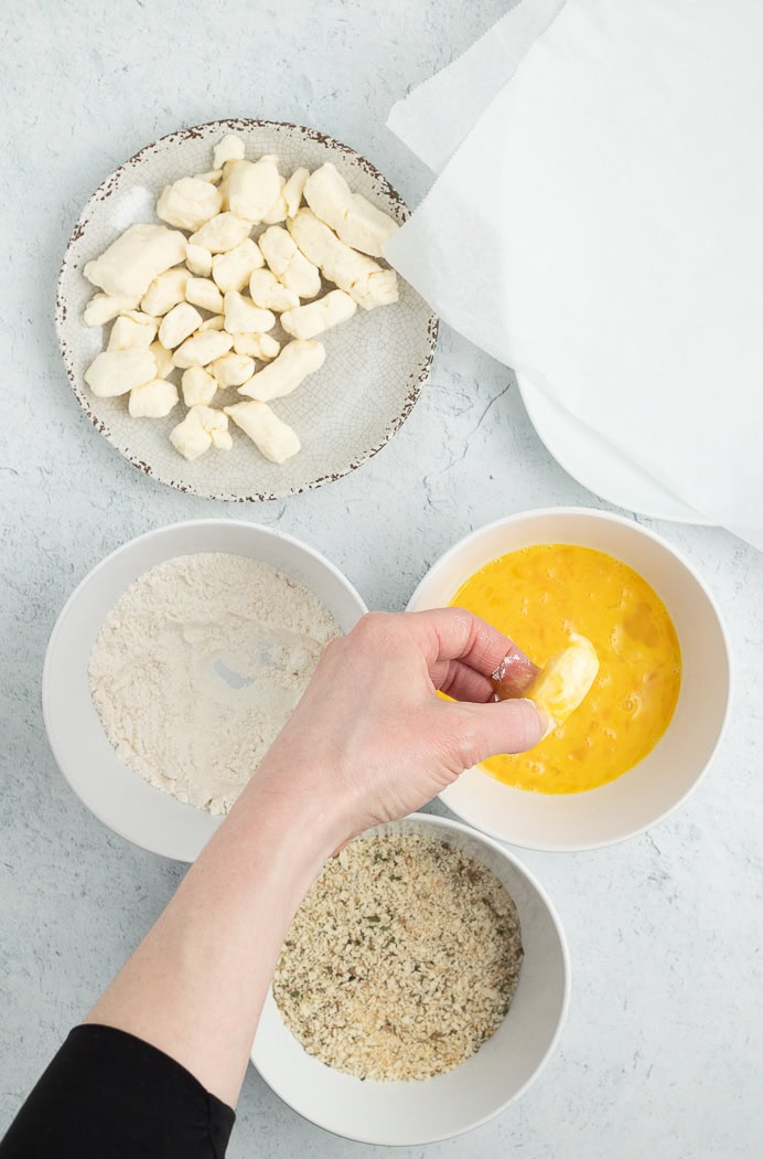 3 bowls of breading and a hand dipping something into a bowl of eggs.