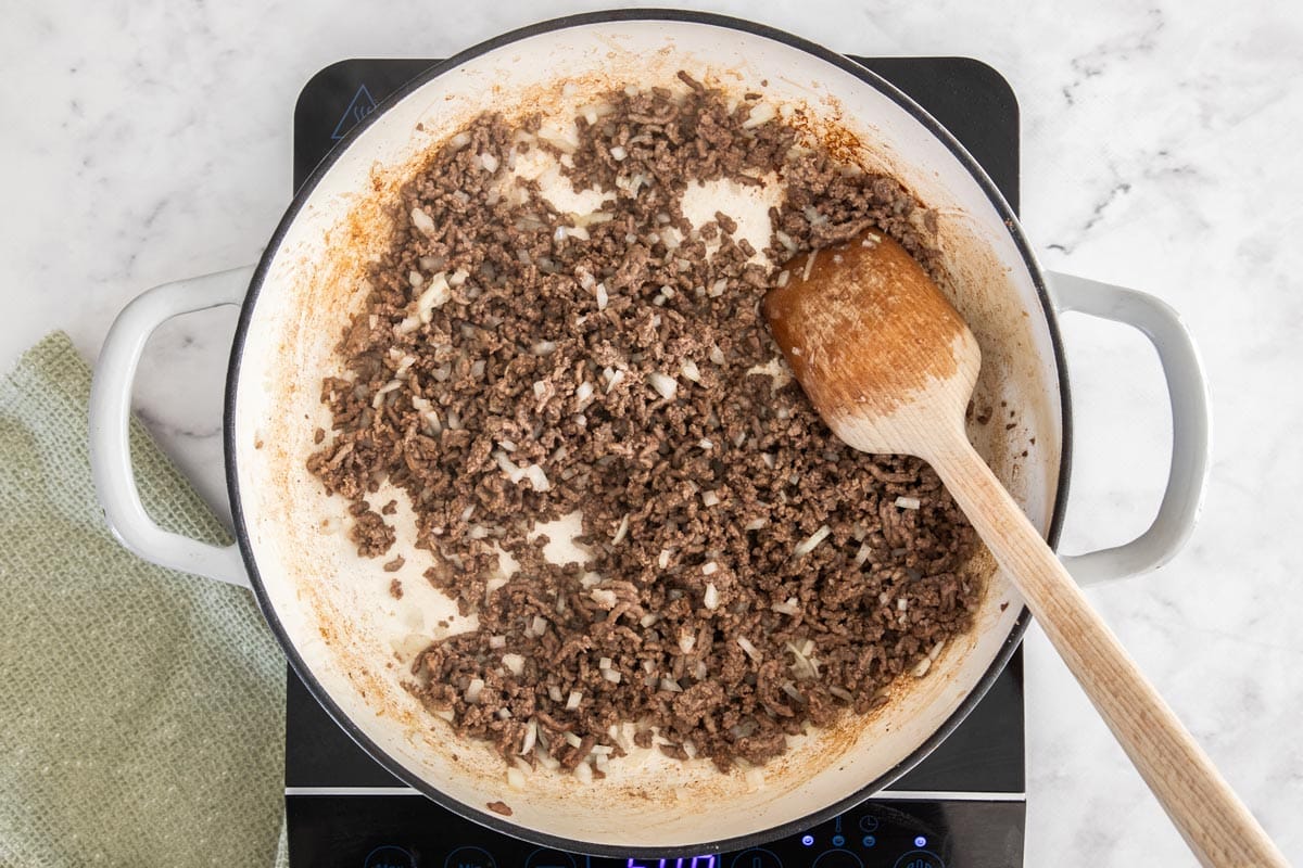 ground chuck and diced onion and garlic in a white skillet with wooden spoon.