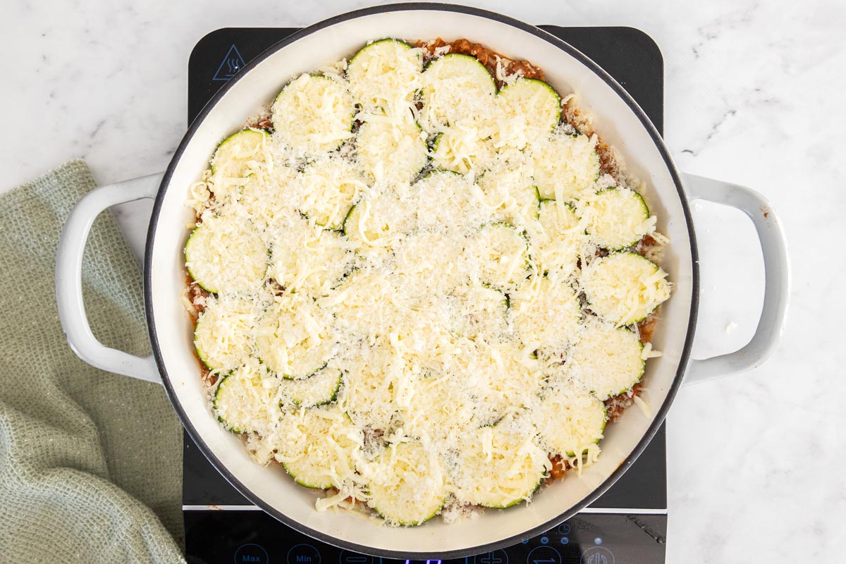 cheese on top of sliced summer squash and maraina in a white skillet.