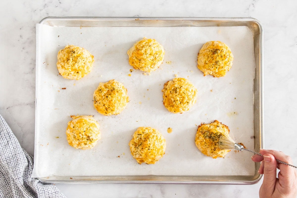 a pastry brush brushing butter on top of rolls on a baking sheet.