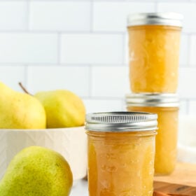 3 jars of pear preserves with fresh pears and cinnamon sticks against a tiled background.