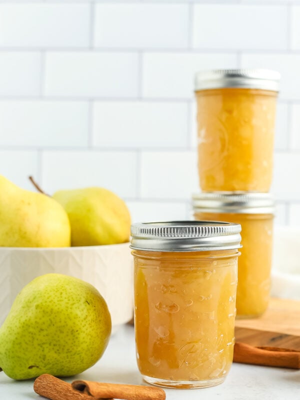 3 jars of pear preserves with fresh pears and cinnamon sticks against a tiled background.