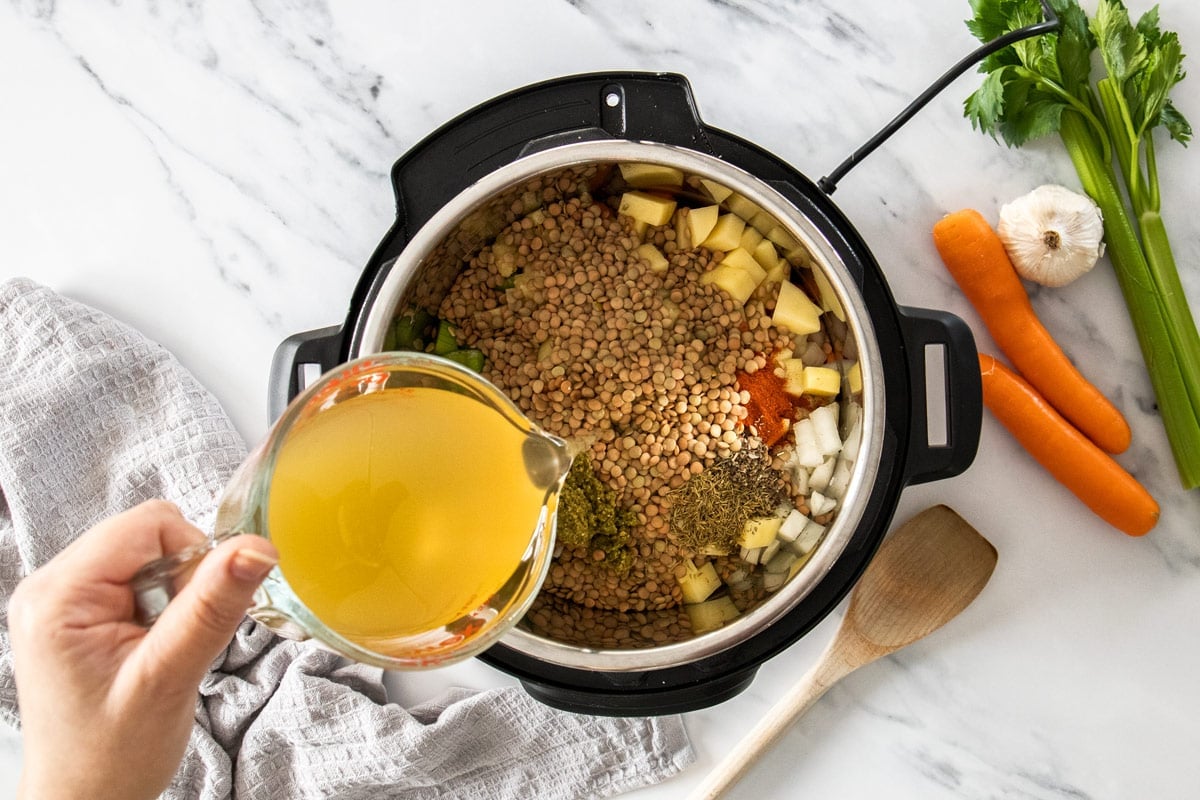uncooked ingredients in a pressure cooker with a measuring cup pouring in chicken broth.