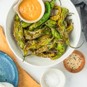 a white bowl of shishito peppers topped with sesame seeds and a small dish of chipotle mayo.