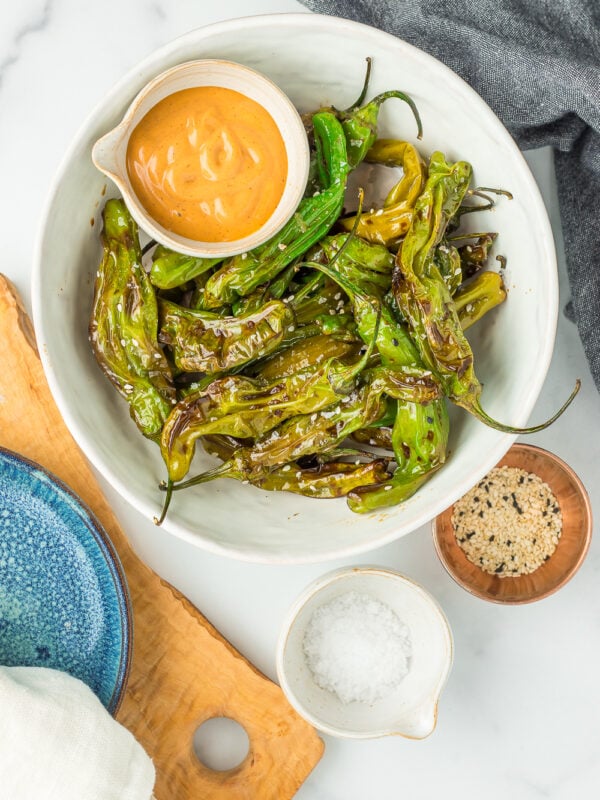 a white bowl of shishito peppers topped with sesame seeds and a small dish of chipotle mayo.