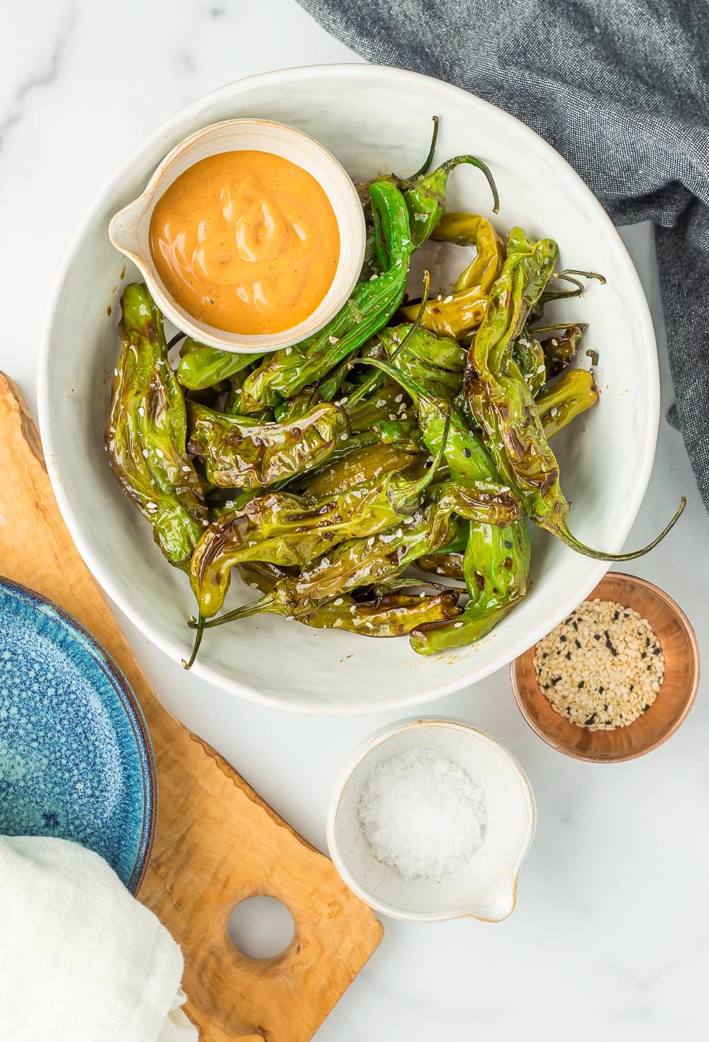 a white bowl of shishito peppers topped with sesame seeds and a small dish of chipotle mayo.