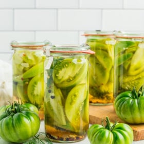 4 jars of pickled green tomatoes with fresh green tomatoes against a tile backsplash.