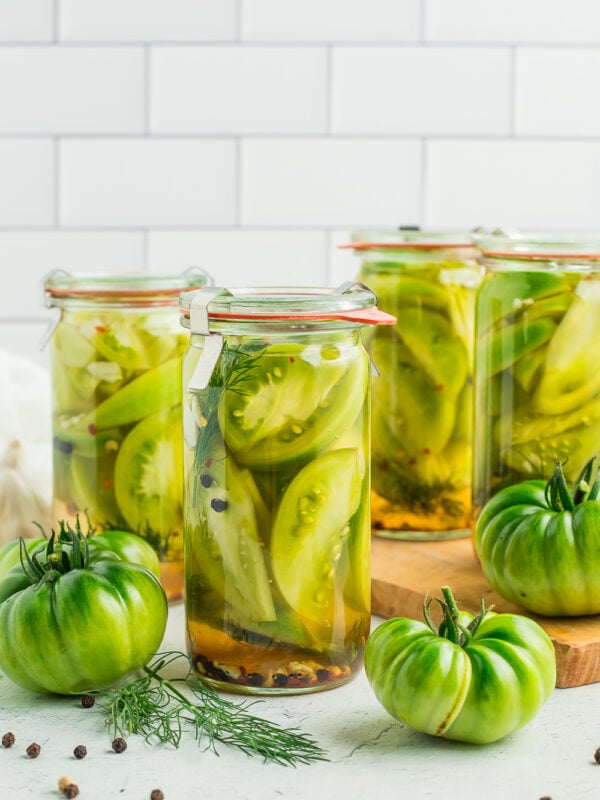 4 jars of pickled green tomatoes with fresh green tomatoes against a tile backsplash.