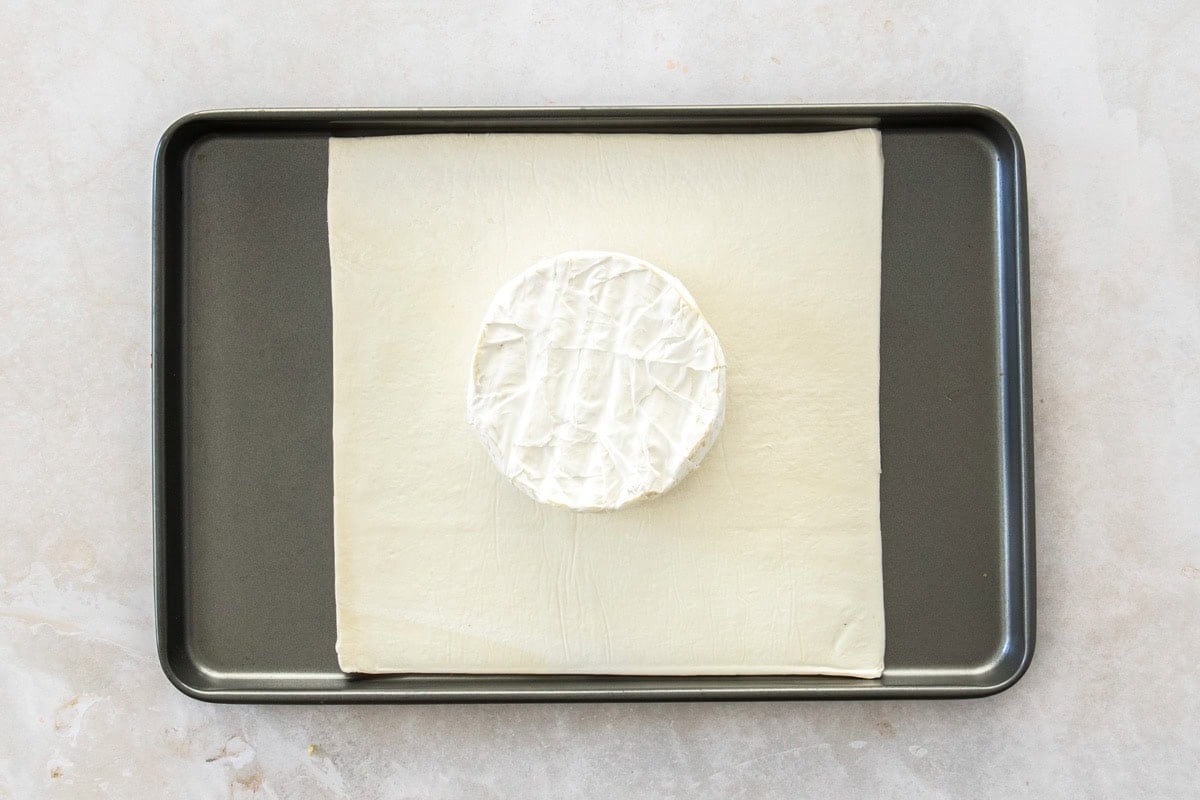 a baking sheet with brie in the middle of a piece of puff pastry.