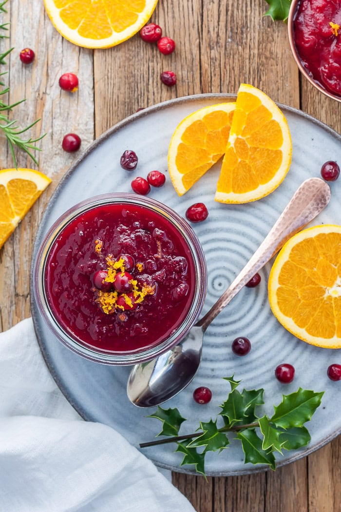 a glass bowl of cranberry sauce on a grey plate with sliced oranges and fresh cranberries.