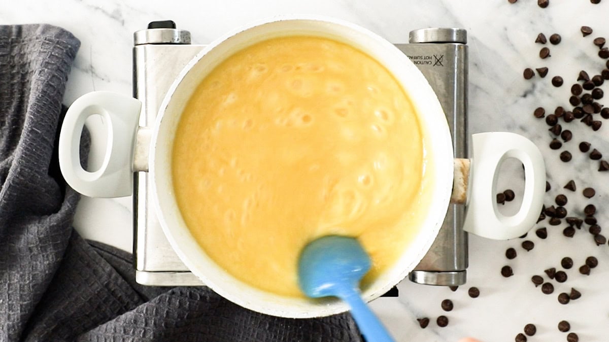 a blue spoon stirring a boiling milk-based sauce in a saucepan.