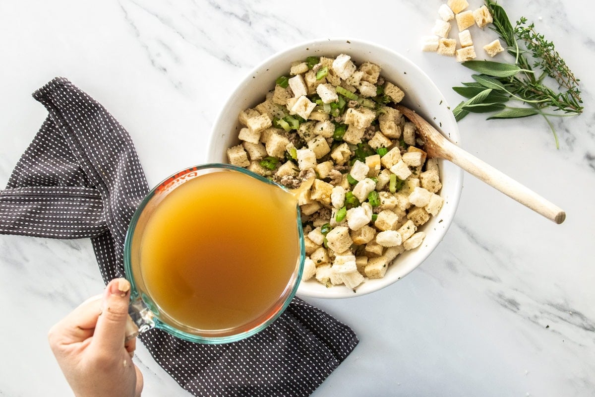 chicken stock being poured over a bowl of bread cubes, celery, and ground sausage.