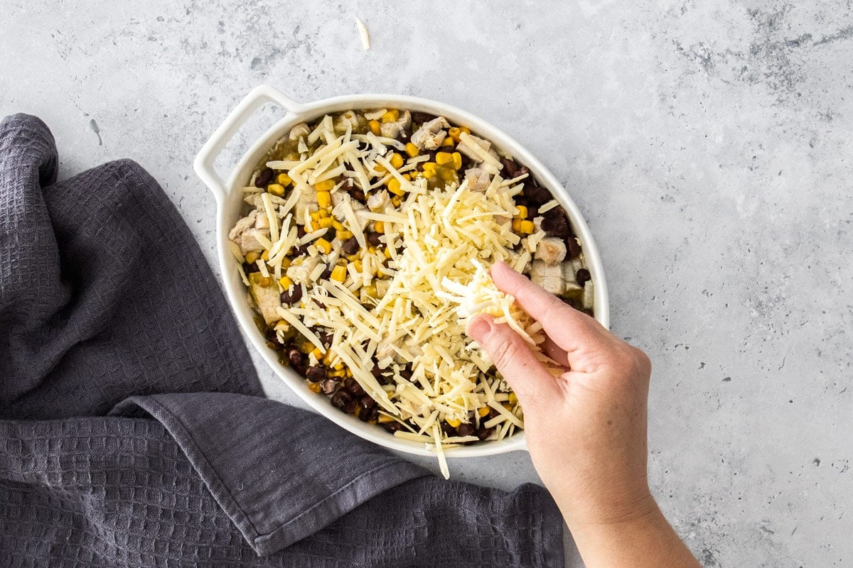 cheddar being sprinkled over ingredients in a white baking dish.