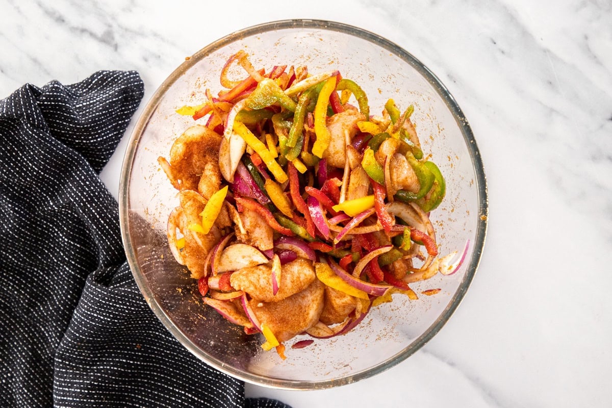 sliced chicken, onions, peppers, and seasoning in a glass bowl.