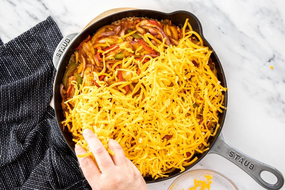 cheese being sprinkled onto the top of a casserole in a cast iron skillet.