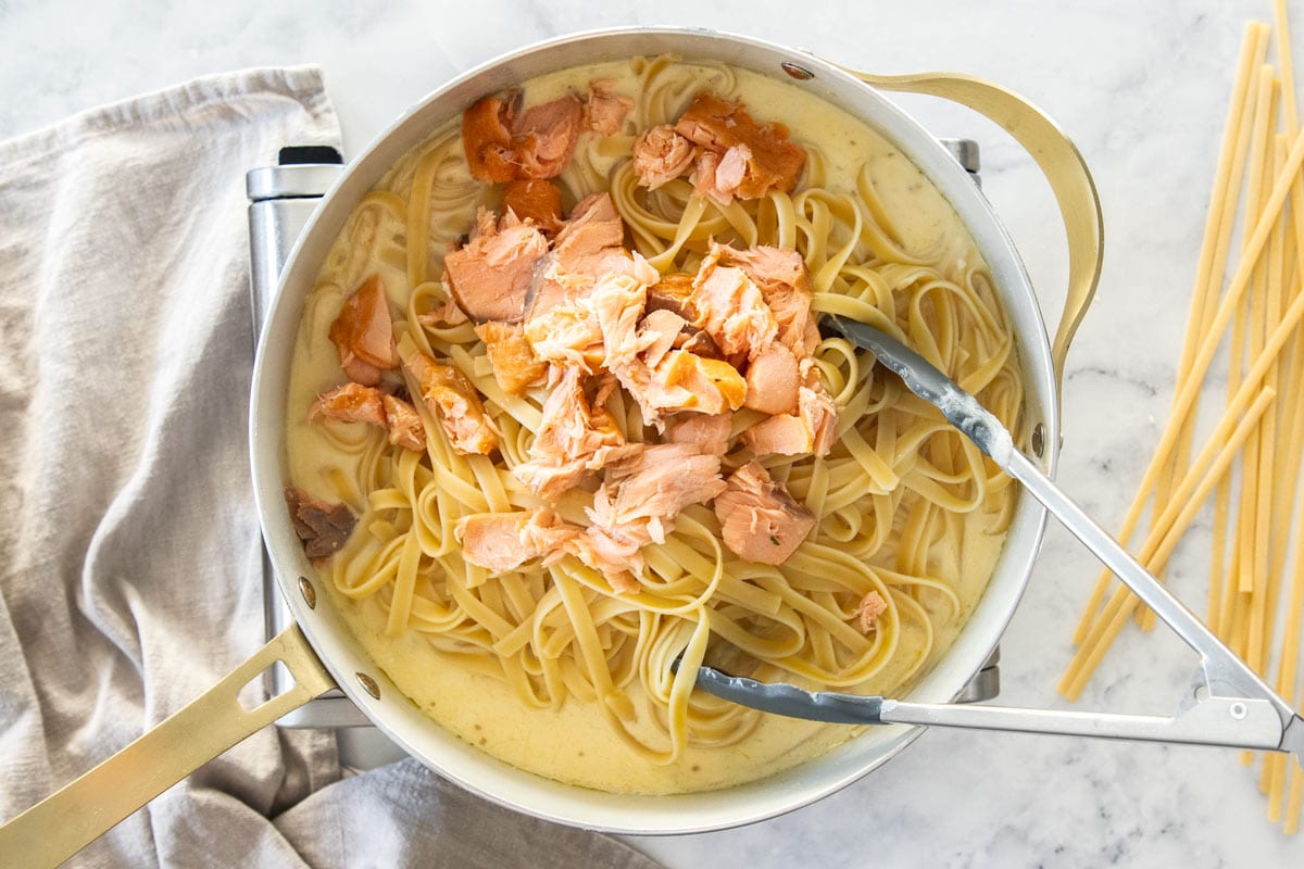 pieces of smoked salmon on top of a white skillet full of noodles and a cream sauce.