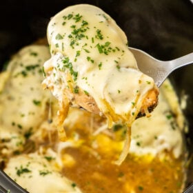 a serving spoon holding a French onion chicken breast topped with cheese above a slow cooker.