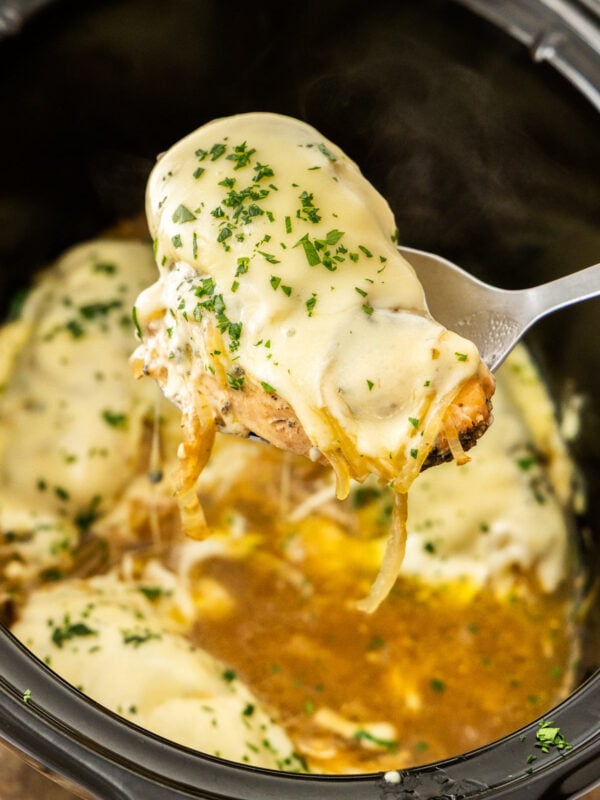 a serving spoon holding a French onion chicken breast topped with cheese above a slow cooker.