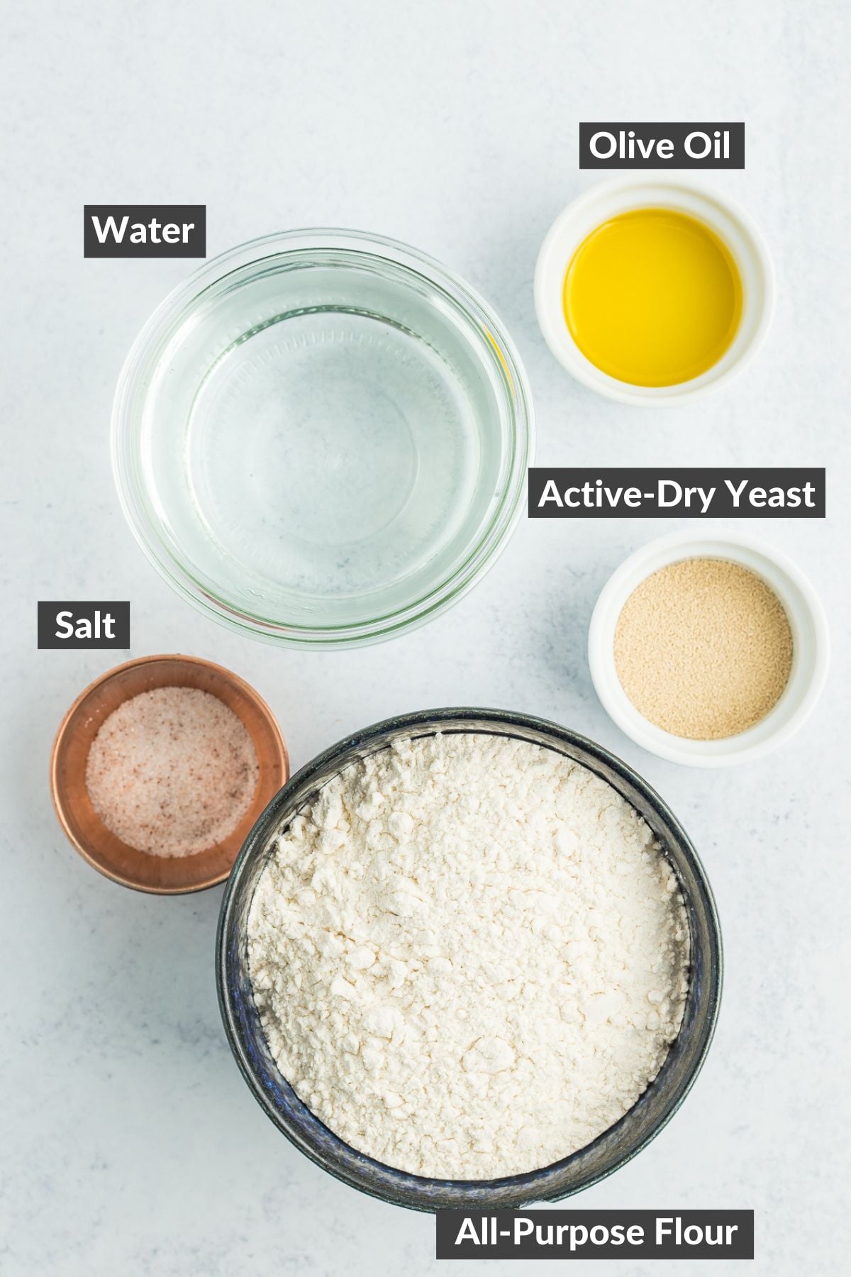 bowls of flour, water, salt, yeast, and olive oil on a grey board.