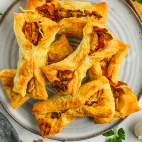 a stack of puff pastry chicken pockets on a grey plate with chopped bacon and shredded Parmesan.