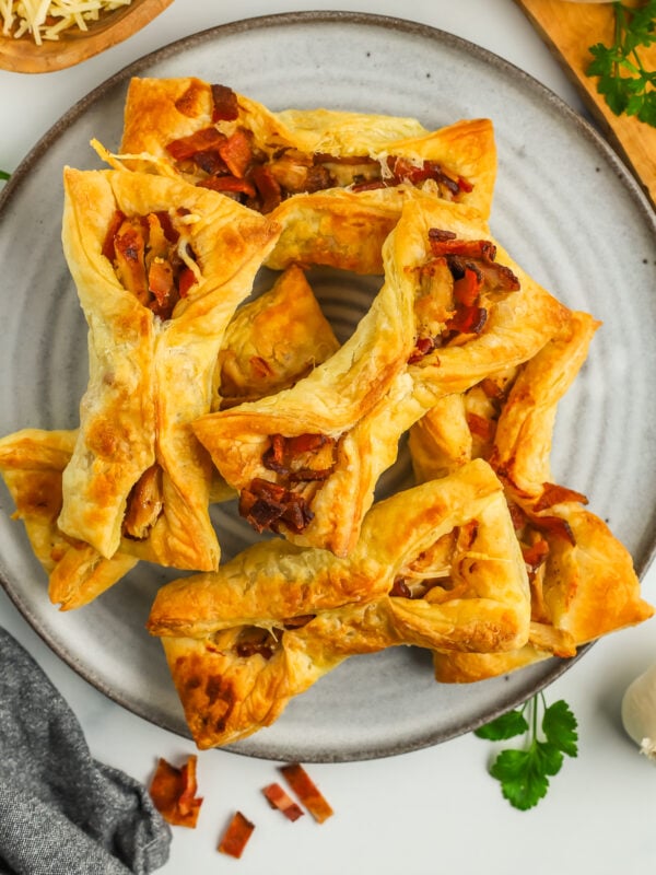 a stack of puff pastry chicken pockets on a grey plate with chopped bacon and shredded Parmesan.