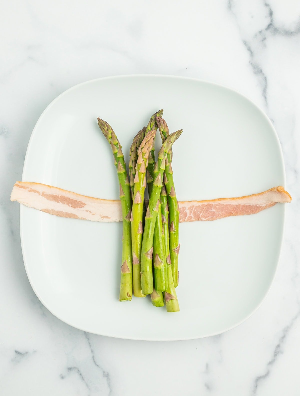 asparagus on top of a long piece of uncooked bacon on a white plate.