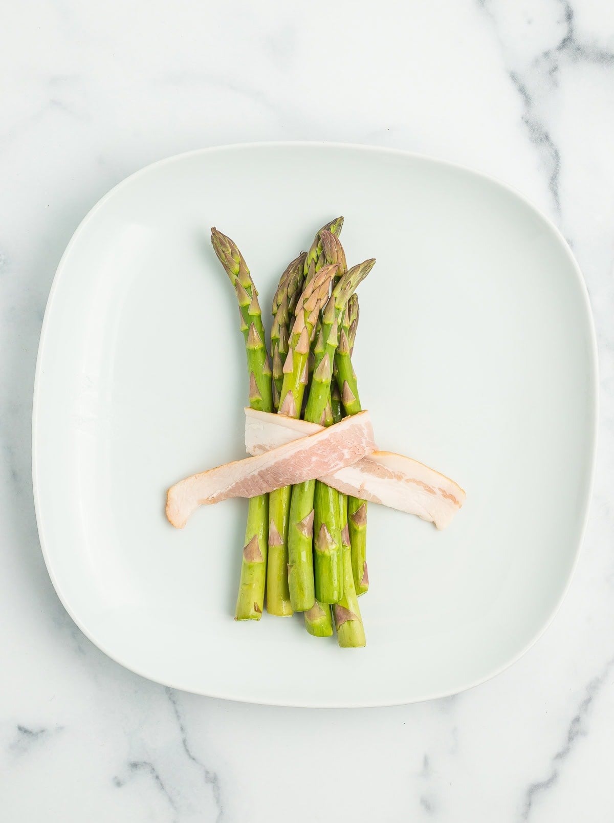 uncooked bacon crossed in front of asparagus bundles on a white plate.