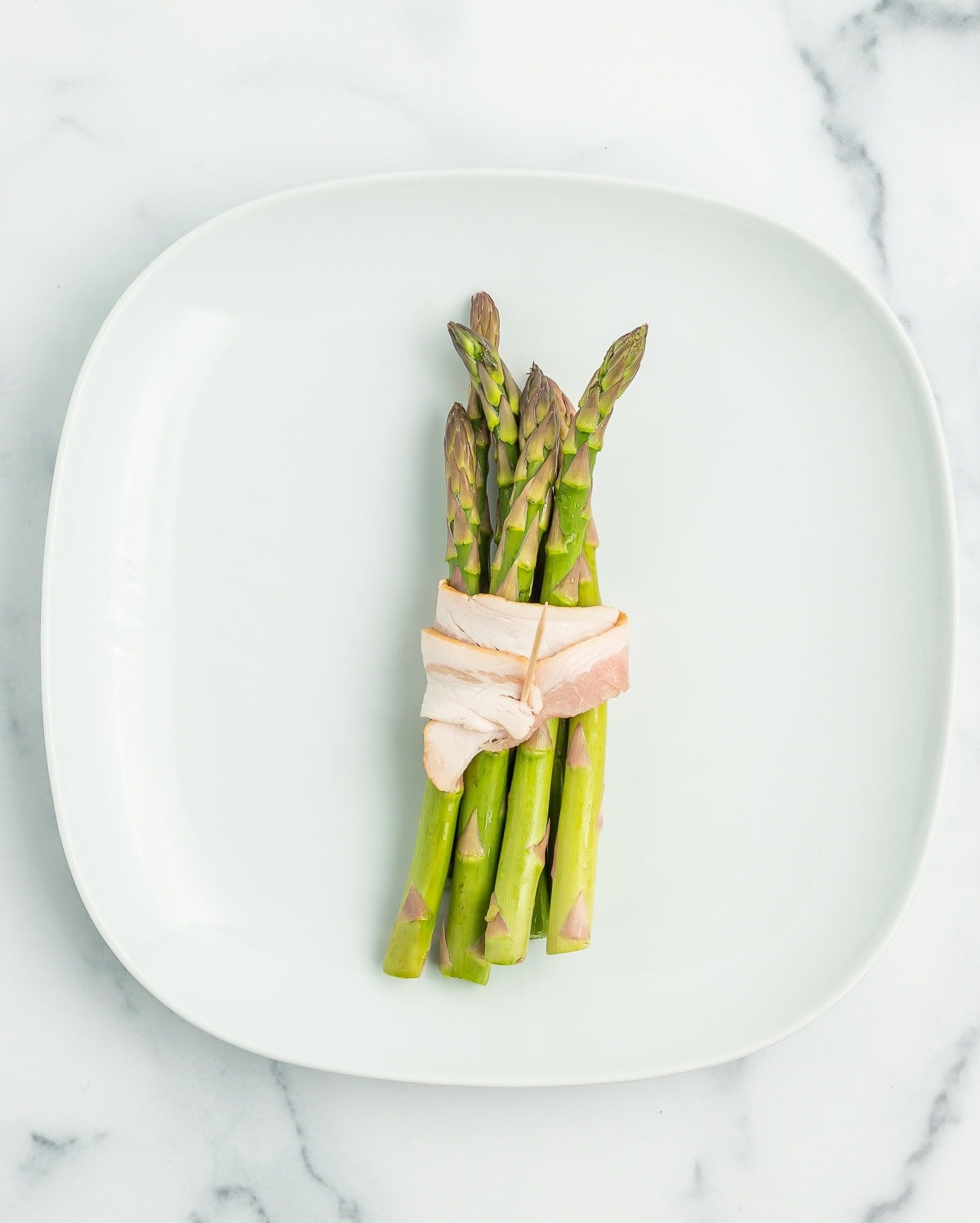 uncooked bacon wrapped around asparagus on a white plate.