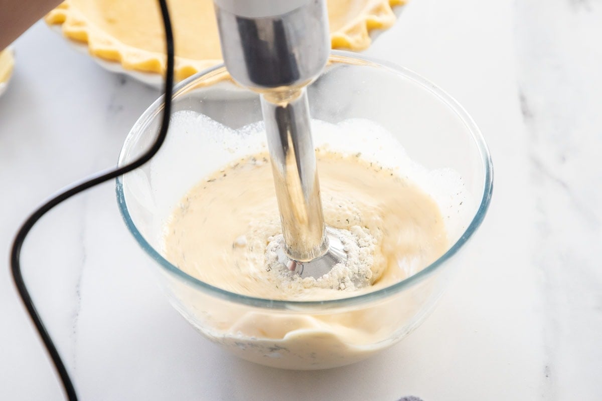 an immersion blender blending eggs, cream, and flour in a glass bowl.