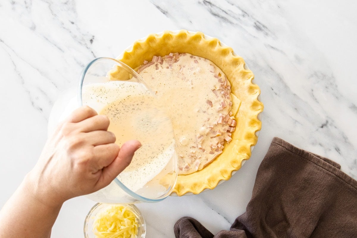 a hand holding a glass bowl pouring eggs and cream into an unbaked pie crust.