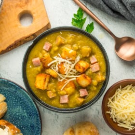 a bowl of slow cooker ham and split pea soup topped with croutons, shredded Parmesan, and baguettes on a grey board.