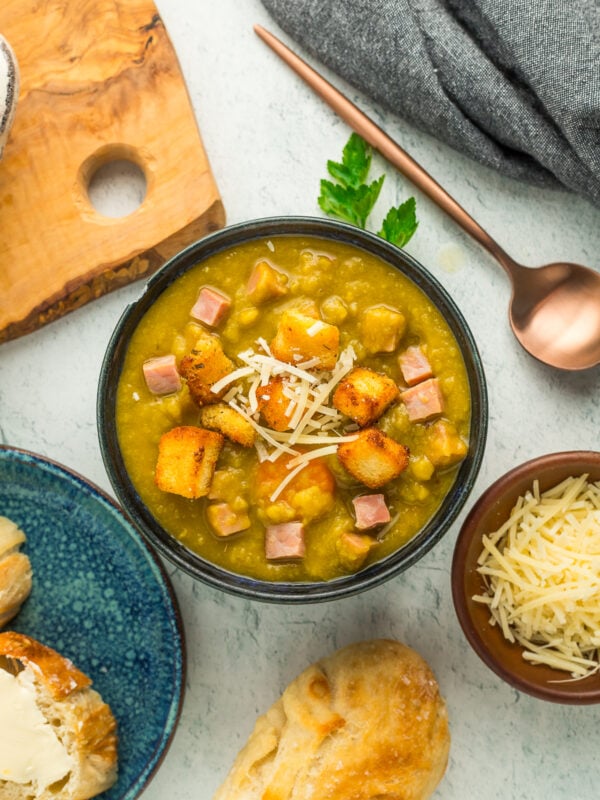 a bowl of slow cooker ham and split pea soup topped with croutons, shredded Parmesan, and baguettes on a grey board.